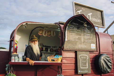 A man with a long white beard and hat stands inside a burgundy food van, with menus displayed outside and snacks, tins, and drinks visible at the serving window.