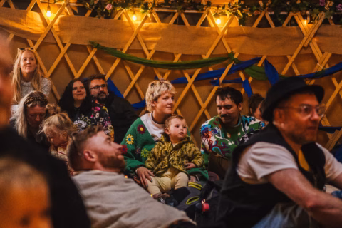 A group of adults and children sit closely together inside a yurt decorated with lights and greenery, watching something off-camera. Some people smile whilst others look attentive.