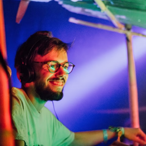 A man with glasses and headphones smiles under colourful lights at what appears to be a music event or party. He is wearing a white T-shirt and has a short beard, with a relaxed and happy expression.