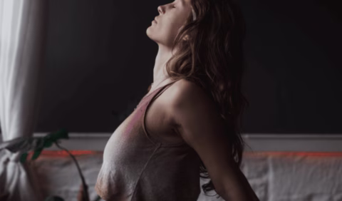 A woman with long hair and a sleeveless top tilts her head back and closes her eyes, appearing relaxed, as sunlight streams through a window nearby.