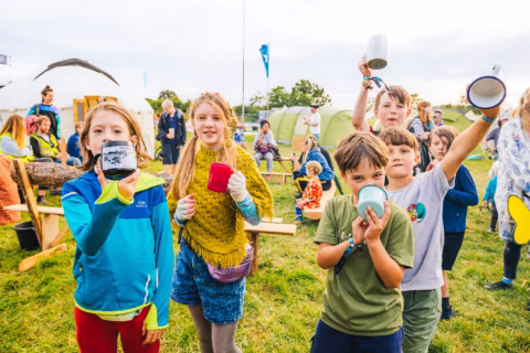 A group of children at an outdoor camp hold up mugs and smile at the camera, with tents and people in the background on a grassy field.