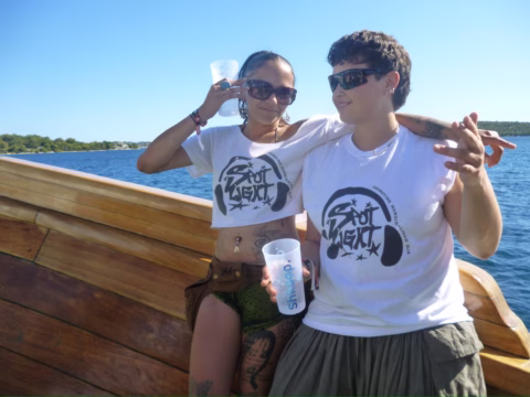 Two people wearing matching Shot Agency T-shirts stand on a wooden boat, smiling and holding drinks, with blue water and a green shoreline visible in the background under a clear sky.