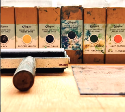 A close-up of a brayer roller and a printing plate on a table, with six boxes of relief inks—black, yellow, blue, green, and orange—lined up in the background.