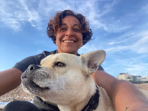 A person with curly hair smiles at the camera while holding a light-coloured dog on a pebble beach, with blue sky and some buildings in the background.