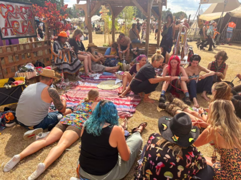 A group of people sit in a circle on blankets outdoors at a festival, some with colourful hair and clothing, chatting and weaving. Tents, trees, and art can be seen in the sunny background.