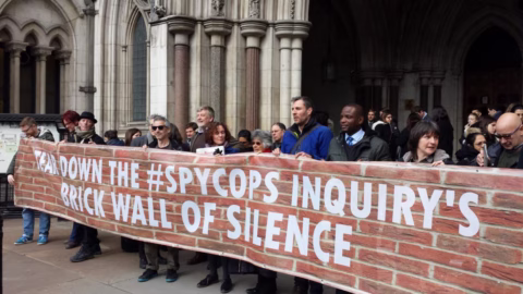 A diverse group of people stand outside a grand stone building holding a large banner that reads “TEAR DOWN THE #SPYCOPS INQUIRY’S BRICK WALL OF SILENCE”.