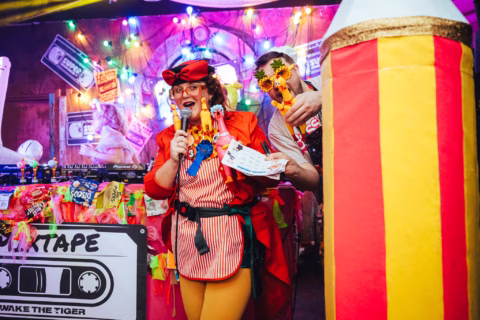 Two people in colourful, playful costumes hold microphones and toys at a decorated stall with cassette tape signs, rainbow lights, and bright decorations, creating a lively and festive atmosphere.