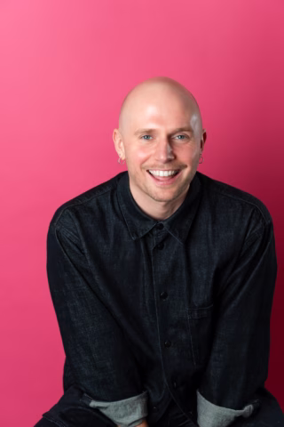 A smiling bald person with light skin wearing a dark denim shirt sits in front of a bright pink background.