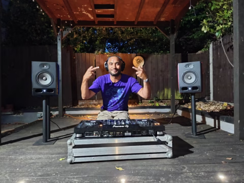A man wearing headphones and a purple shirt poses with peace signs behind DJ equipment on a wooden patio, flanked by large speakers, under a pergola with outdoor lighting.