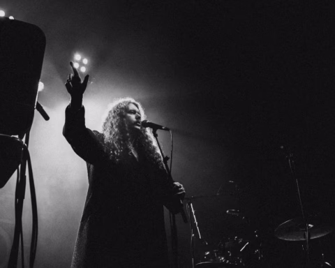 A person with long curly hair sings into a microphone on a dimly lit stage, raising one hand dramatically. Stage lights shine behind, casting a moody, artistic atmosphere in black and white.