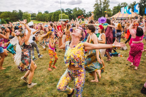 A large group of people in colourful, festive outfits dance joyfully outdoors on grass at a lively festival, with tents and trees in the background under a partly cloudy sky.