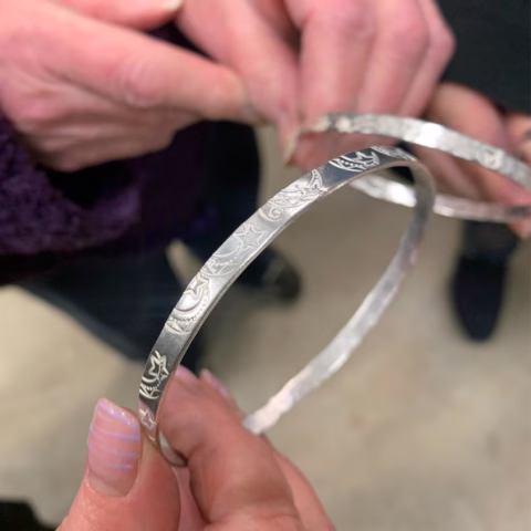 Two people hold engraved silver bangles. One person’s hand, with purple striped nails, holds a bangle close to the camera, revealing detailed patterns and stars on its surface. Another hand holds a second bangle in the background.