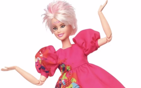 A doll with short, spiky pink and white hair wears a bright pink dress with puffed sleeves and colourful abstract patterns, smiling with raised arms against a white background.