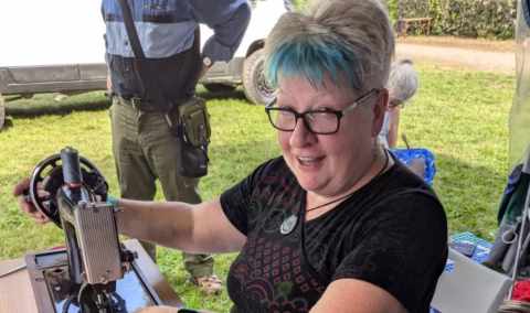 A smiling woman with short grey and blue hair uses a sewing machine outdoors. She wears glasses and a patterned shirt. A person stands behind her and a van is visible in the background.
