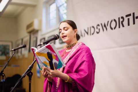 A woman in a bright pink traditional outfit reads from a colourful book at a microphone stand, speaking in front of a banner that partially reads WANDSWORTH.