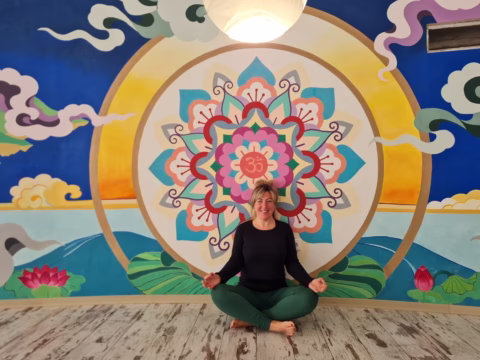 A woman sits cross-legged and smiling on a wooden floor in front of a colourful mandala mural with cloud, sun, and lotus designs. She wears a black top and green trousers, hands resting on her knees in a meditative pose.