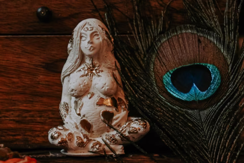 A white, ornate figurine of a pregnant woman in a meditative pose sits beside a vibrant peacock feather, both placed on a dark wooden surface. The figurine has gold accents and closed eyes.