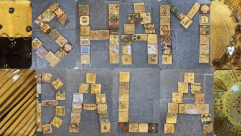 Tiles arranged on a stone surface spell out SHAMBALA. Each tile features unique patterns, symbols, or artwork, mostly in shades of yellow, brown, and gold. Some larger, decorated tiles frame the words.