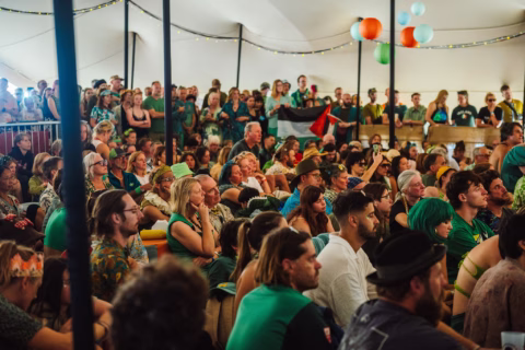 A large, diverse crowd sits closely together inside a marquee, attentively watching an event. Colourful lanterns hang above, and some people wear bright clothing and costumes. A few hold up a flag at the back.