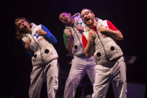 Three people wearing white puffy waistcoats and white trousers perform energetically on stage, singing with expressive faces and gestures. The background is dark, highlighting the performers under stage lighting.