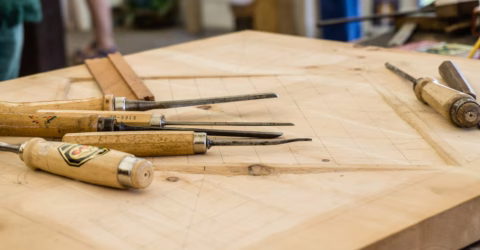 Several wood carving chisels with wooden handles are laid out on a large wooden workbench, which has grid lines and measurements marked on its surface.