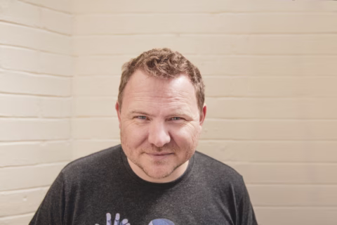 A man with short, curly brown hair and a trimmed beard is standing in front of a cream-coloured brick wall, wearing a dark T-shirt and smiling slightly at the camera.