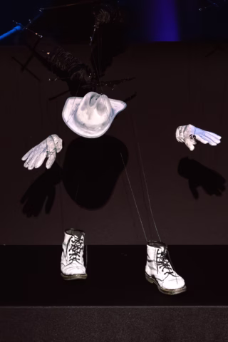 A marionette costume featuring a white cowboy hat, white gloves, and white boots is suspended by strings, with no visible body, set against a dark background.
