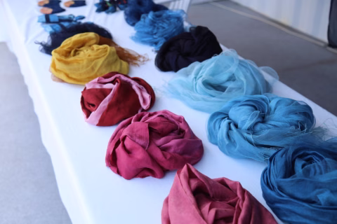 Colourful scarves are neatly displayed on a white table. The scarves are rolled or loosely folded in shades of red, blue, yellow, black, and teal, arranged in rows on the tabletop.