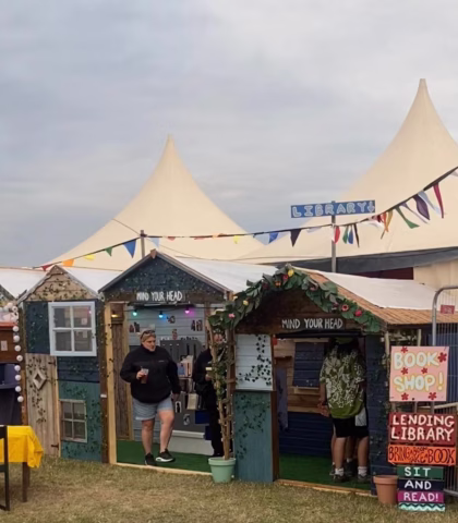 Two people stand outside a small, colourful pop-up library and bookshop at an outdoor event, with bunting, “Mind Your Head” and “Library” signs, and large white marquees in the background.