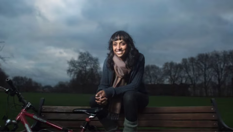 A woman with dark hair sits on a park bench, smiling at the camera. She is wearing a scarf and jumper, with a red bicycle beside her. The sky is cloudy and trees are visible in the background.