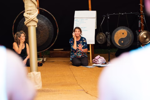 A woman sits cross-legged on the floor, gesturing with her hands and smiling, while another woman sits nearby. Behind them are large gongs, a chart with notes, and some fabric, creating a relaxed, workshop-like atmosphere.