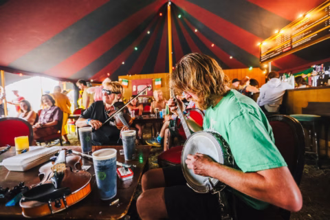 Two people play a banjo and a violin inside a colourful tent with fairy lights. Others sit around, chatting and enjoying drinks at tables in a lively, casual atmosphere.