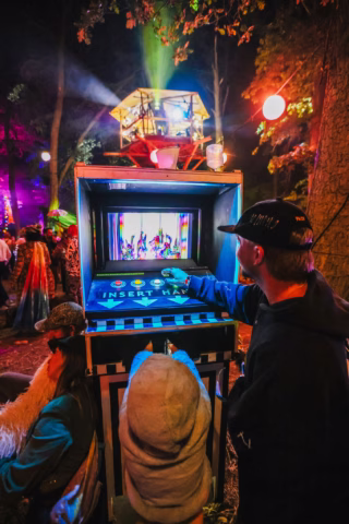 Three people interact with a large, colourful arcade machine outdoors at night, surrounded by trees and vibrant lights, with costumed figures and a stage in the background.