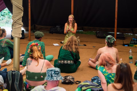 A woman sits cross-legged speaking to an attentive group indoors. The audience, mostly dressed in green, sits on the floor facing her. Natural light comes from outside the marquee-like structure.