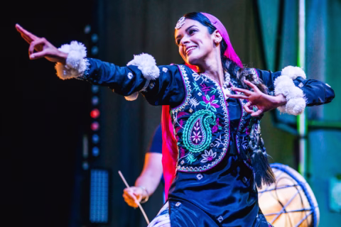 A dancer performs energetically in traditional, colourful attire with intricate embroidery, smiling and posing with one arm extended. Bright stage lighting highlights the vibrant details of the costume.
