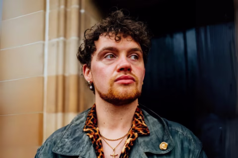 A person with curly hair, a beard, and moustache stands outside near a stone wall, wearing a green jacket with a gold pin, a leopard print shirt, and several necklaces and earrings, looking off to the side.