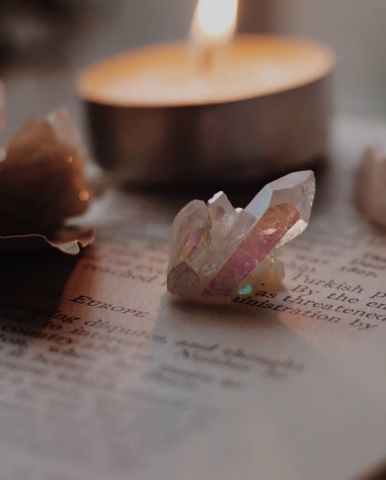 A cluster of clear crystals with rainbow reflections rests on an open book, with a lit tealight candle softly glowing in the blurred background.