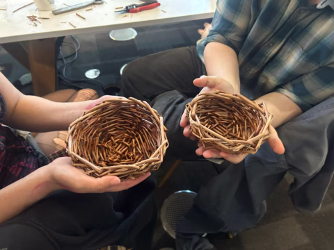 Two people sit at a table, each holding a small, handwoven wicker basket. Basket-making materials and tools are visible on the table in the background.