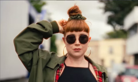 A woman with red hair in a top bun, wearing round black sunglasses, gold hoop earrings, an olive green jacket, and a black top, stands outdoors with one hand on her head, looking confidently at the camera.