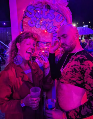 Two people pose playfully under purple lights at night. The person on the left laughs, holding a drink and wearing spiral earrings, whilst the person on the right touches their head, exposing their midriff. A colourful sign reads Satsuma Listen behind them.
