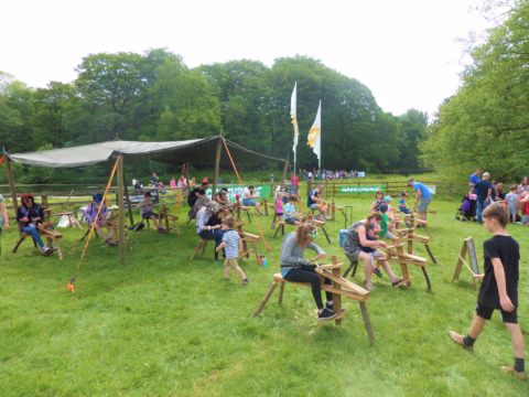 Children and adults sit on wooden benches and work on craft activities under a canopy in a grassy outdoor area, surrounded by trees and more people in the background.