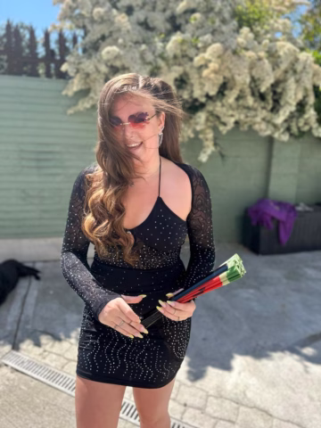 A woman in a sparkly black dress and red sunglasses smiles outdoors, holding three roses. Her long hair is blowing in the wind, and there are white flowering bushes and a green fence in the background.