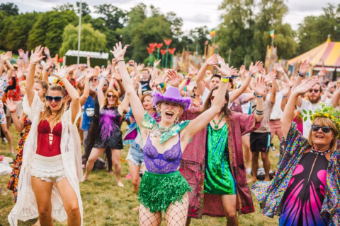 A lively crowd of people in colourful, festive costumes dance with their hands raised outdoors at a summer festival, with trees and tents visible in the background.