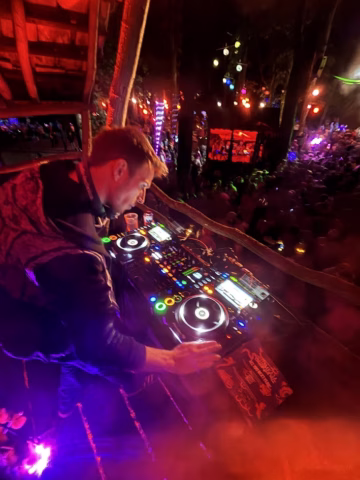 A DJ mixes music on a colourful control board at an outdoor night event, with a lively crowd and vibrant lights in the background among trees.