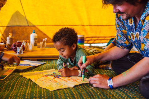 A young child and an adult lie on a mat under a yellow tent, colouring together on a large piece of paper. Art supplies like felt-tips, pencils, and glue are scattered around them, creating a cosy and creative scene.