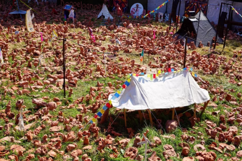 A white marquee with colourful flags stands among hundreds of small terracotta figures scattered on grass, with other marquees and banners visible in the background.