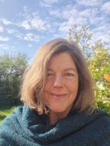 A woman with shoulder-length light brown hair smiles outdoors, wearing a teal shawl. Behind her are green trees, flowering shrubs, and a blue sky scattered with clouds.