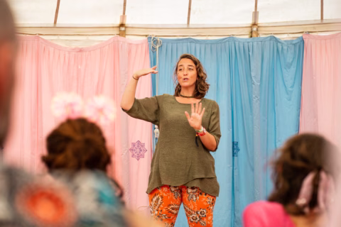 A woman stands in front of pink and blue curtains, gesturing with her hands as she speaks to a seated audience. She wears a green top and orange patterned trousers.