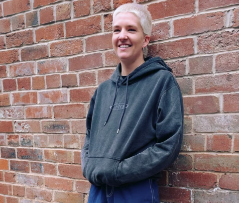 A person with short blonde hair wearing a dark hoodie and blue trousers stands smiling in front of a red brick wall.