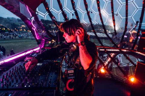 A DJ wearing headphones and a staff badge mixes music at a console inside a futuristic, dome-like booth with colourful lights, whilst a festival crowd gathers outside in the evening.